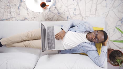 Afrikanischer-Mann-Liegt-Zu-Hause-Auf-Dem-Sofa,-Ruht-Sich-Aus-Und-Benutzt-Laptop.